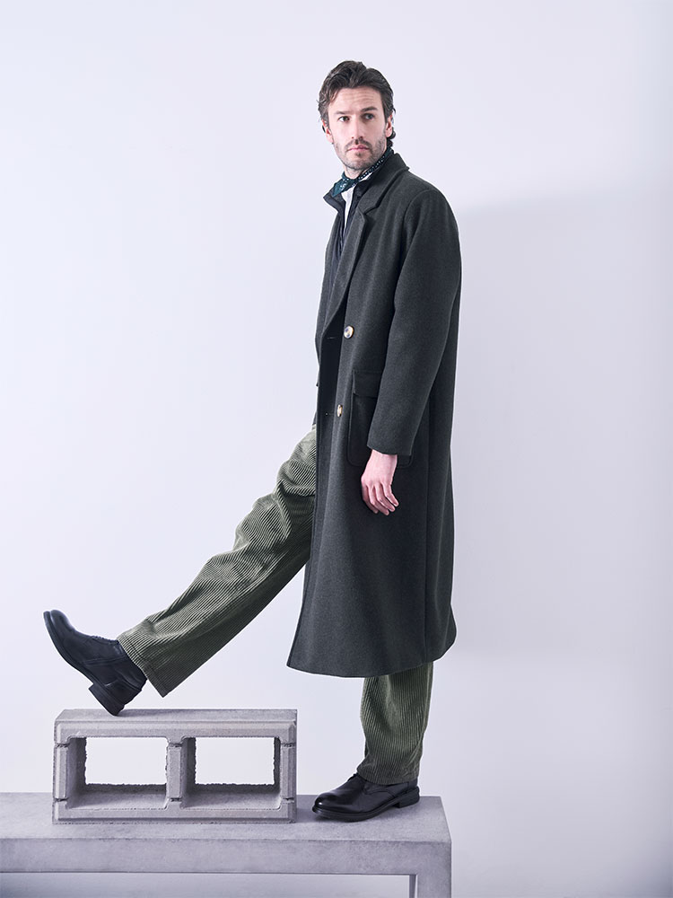 Ein Mann in einem langen dunkelgrünen Mantel, einer grünen Hose und schwarzen Schuhen steht mit einem Fuß auf Betonblöcken und mit dem anderen auf dem Boden und blickt vor einem schlichten hellen Hintergrund zur Seite.